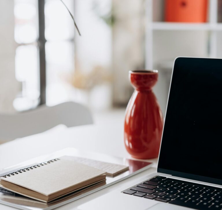 A minimalist home office setup featuring a laptop, notebook, and a red vase on a white desk.