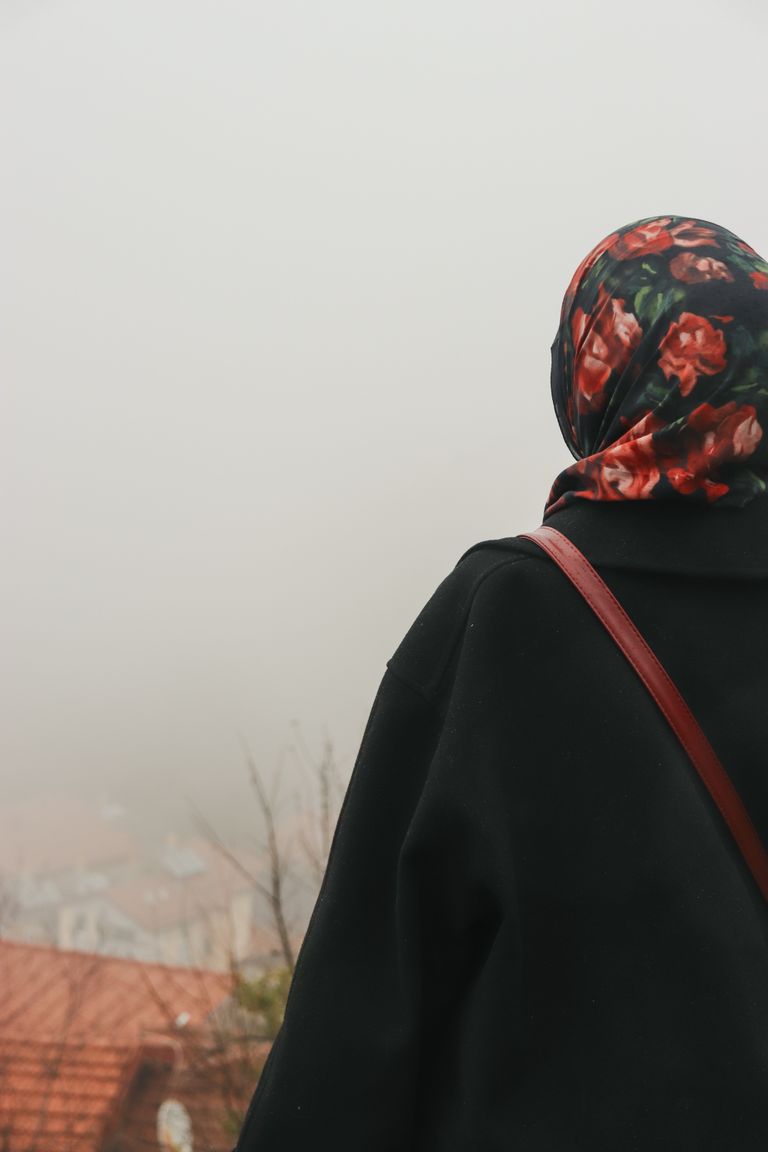 A woman in a floral hijab standing outdoors, looking at a foggy cityscape. Captured from behind.