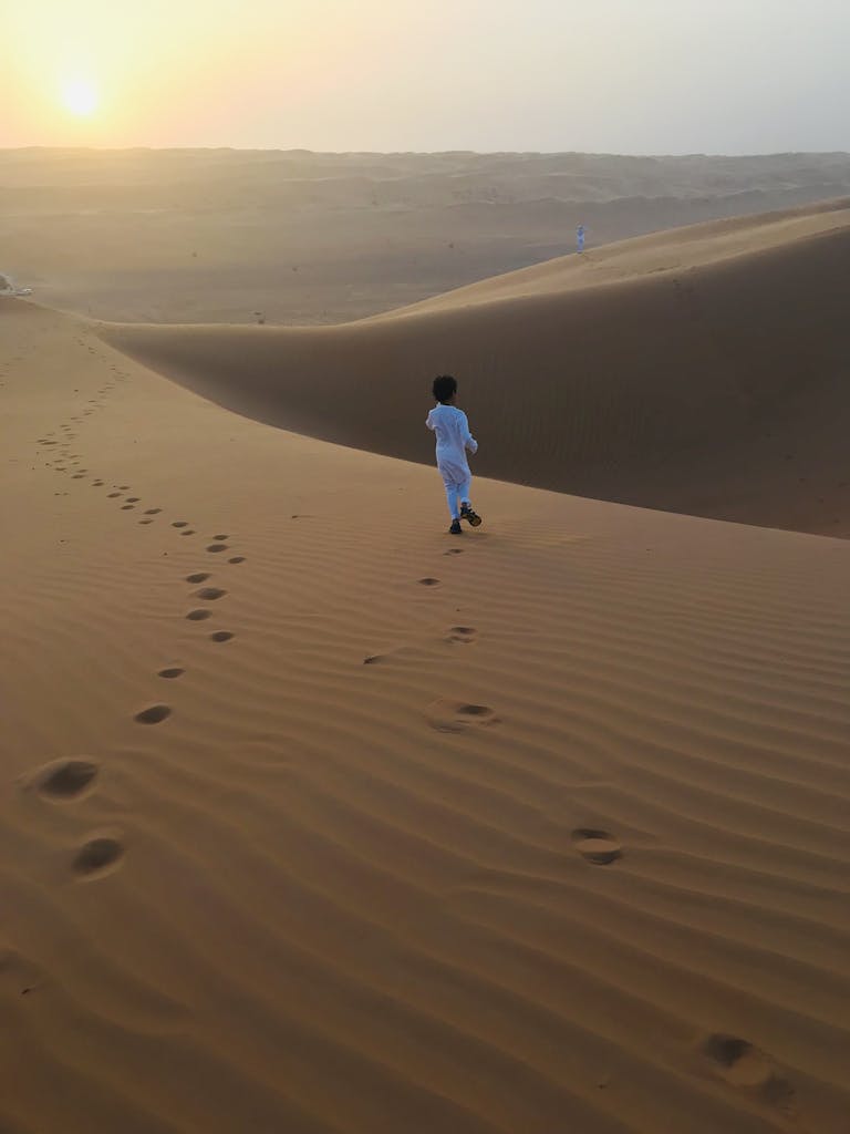 A young child in traditional attire walks on sand dunes during a warm sunset.
