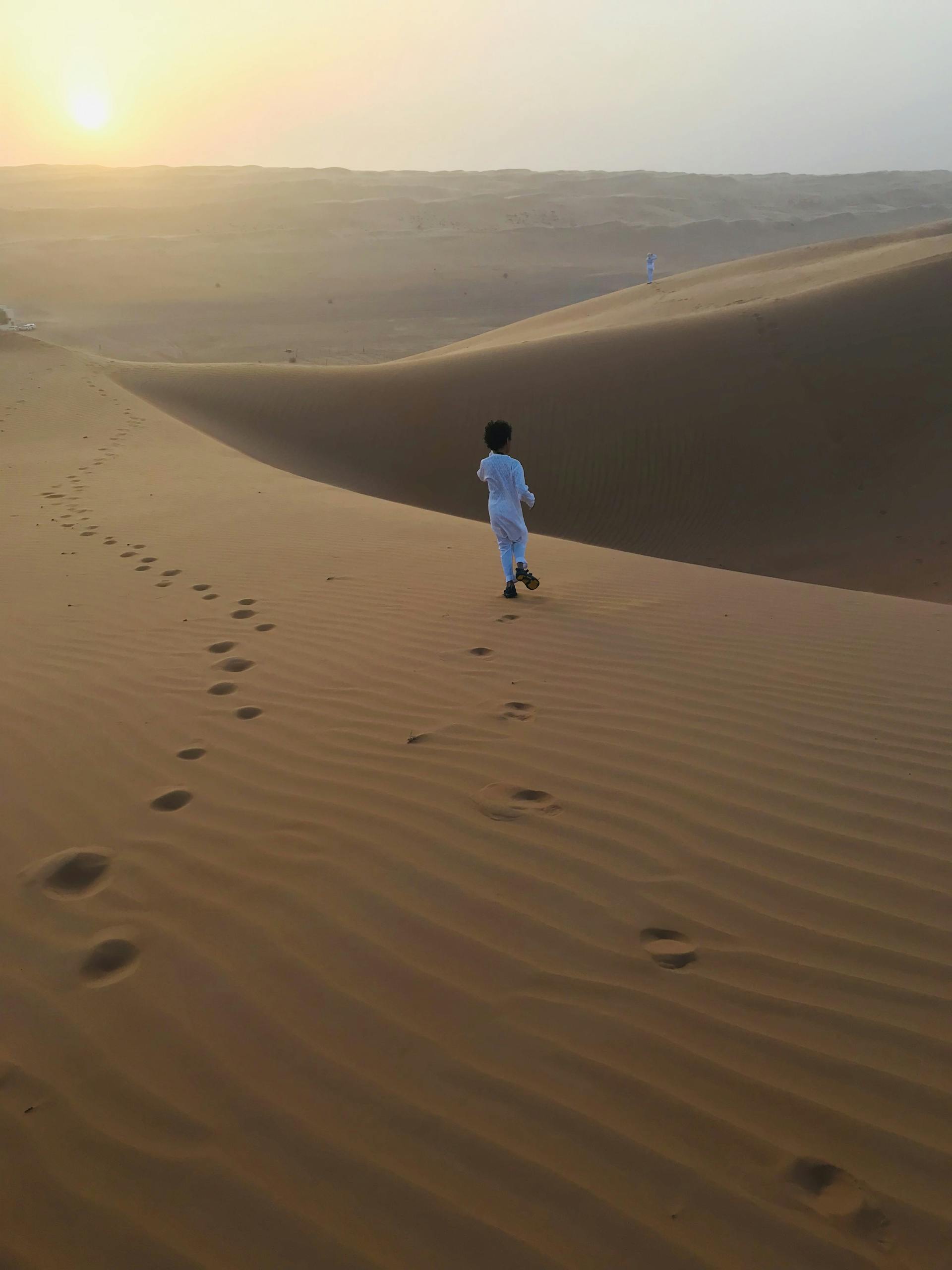 A young child in traditional attire walks on sand dunes during a warm sunset.