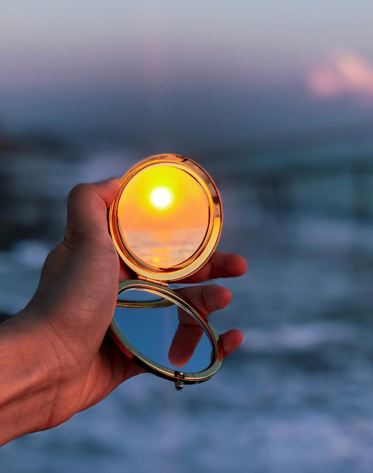 Hand holds mirror reflecting a vibrant sunset on a scenic beach.
