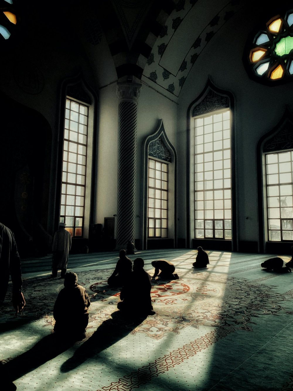 Silhouetted individuals praying inside a sunlit mosque with intricate architecture.